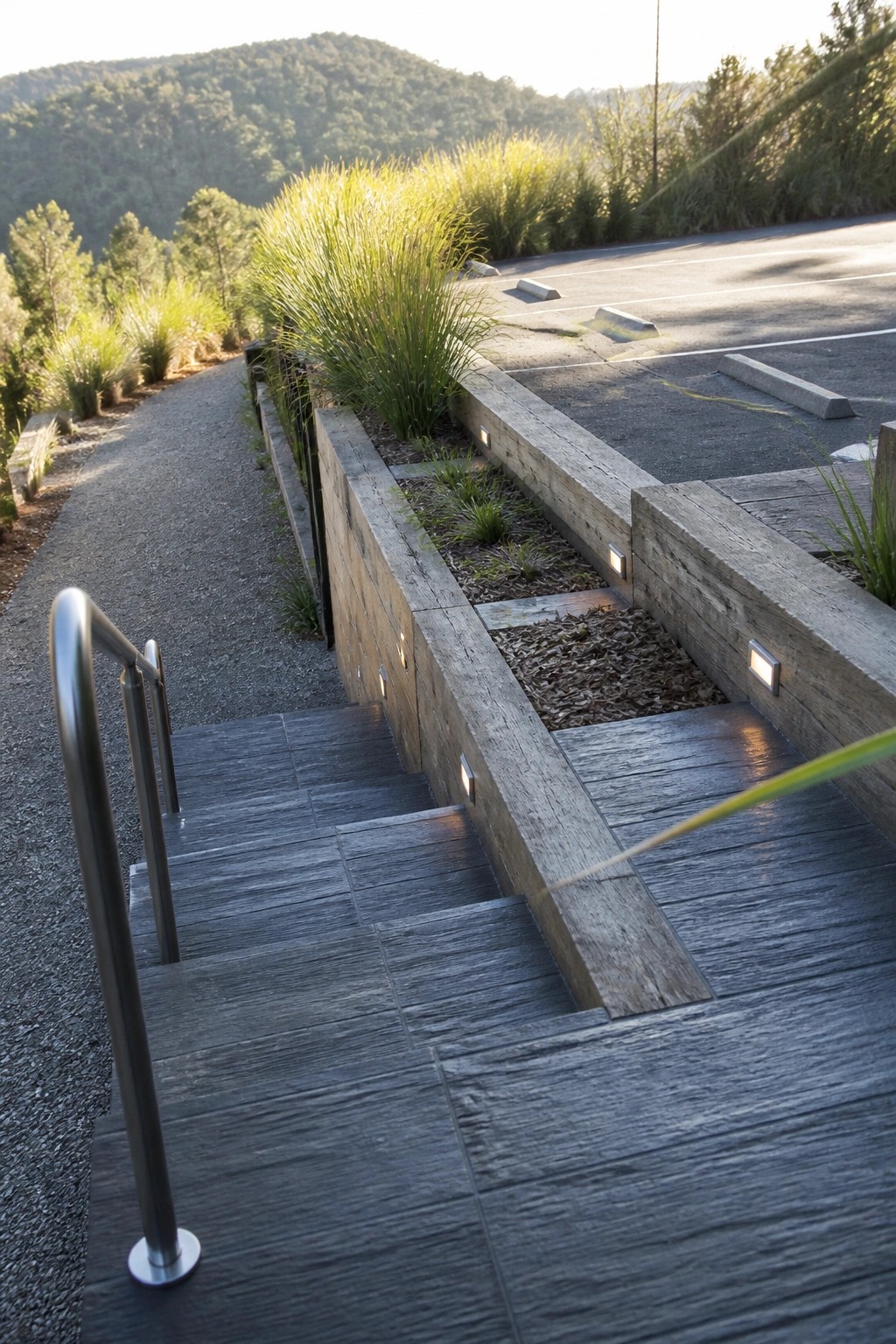 Dark textured tiles form a set of outdoor stairs with a metal handrail, bordered by wooden retaining walls with small lights and a gravel path beside a paved parking area.