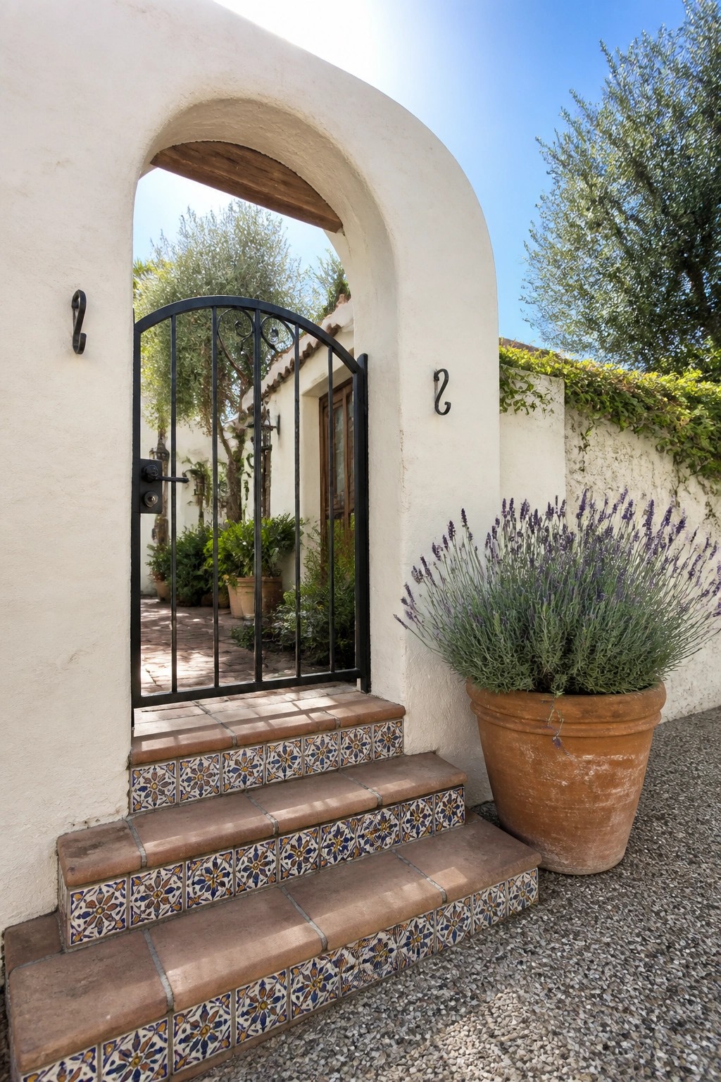 A white stucco house entrance with an arched opening, black metal gate, tiled steps, and a large terracotta pot with lavender nearby.