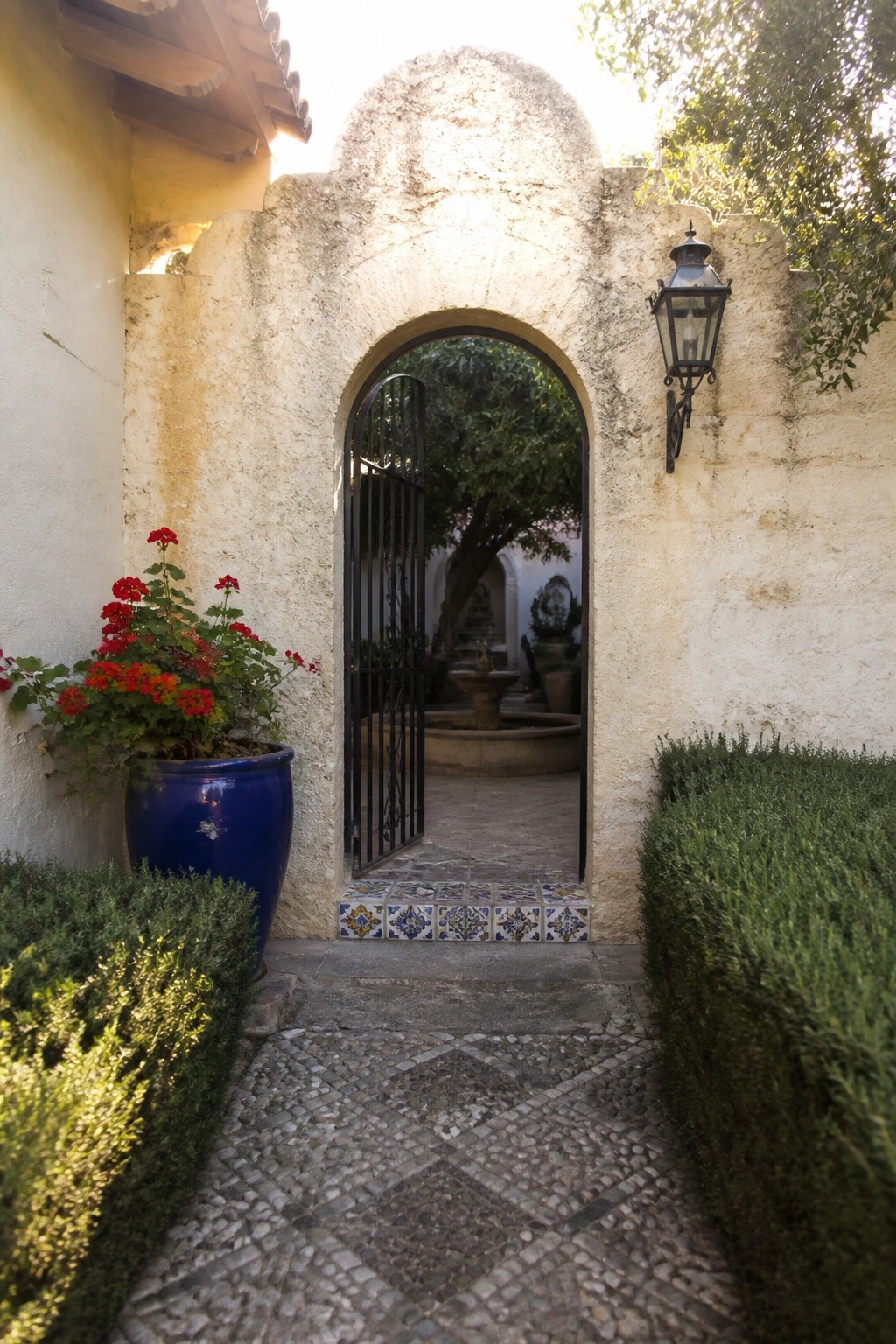 A stone arch with a black metal gate stands open above a tiled threshold, leading into a courtyard with a fountain.