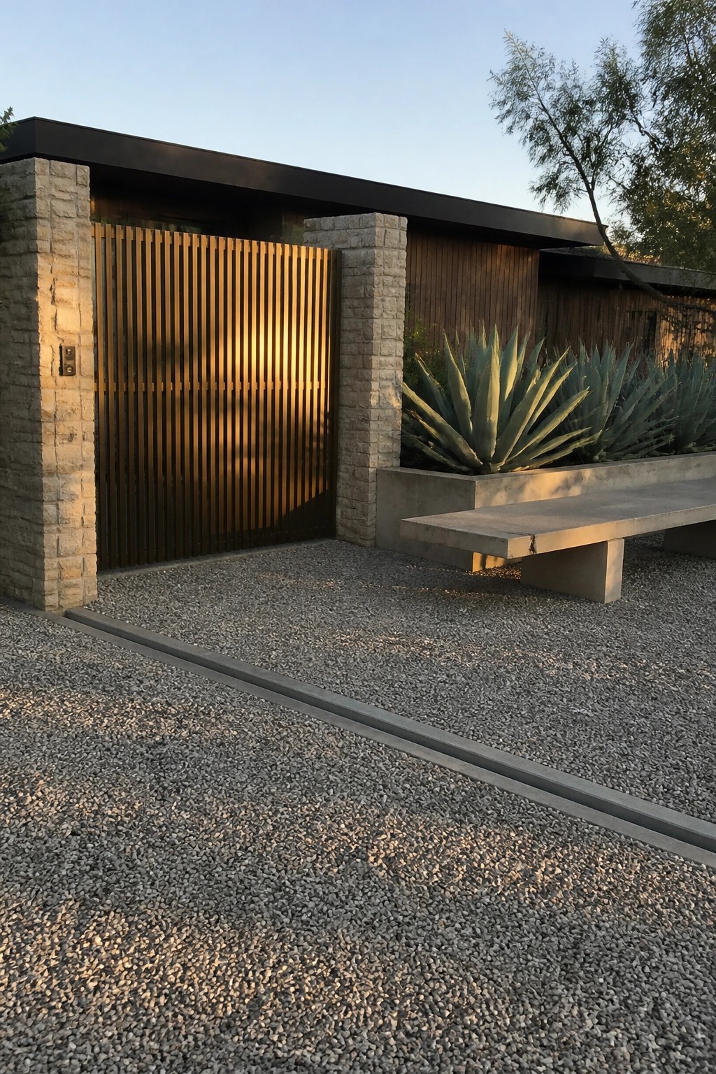 A modern main gate with vertical wooden slats between stone pillars, next to a gravel driveway and concrete bench with agave plants.