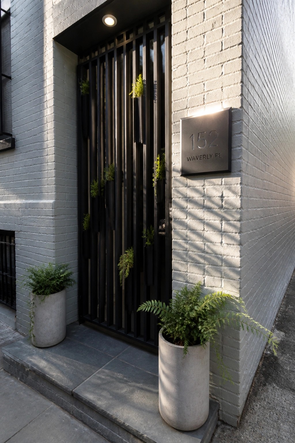 A black iron gate with vertical bars on a white brick wall next to a metal plaque and two concrete planters with ferns.