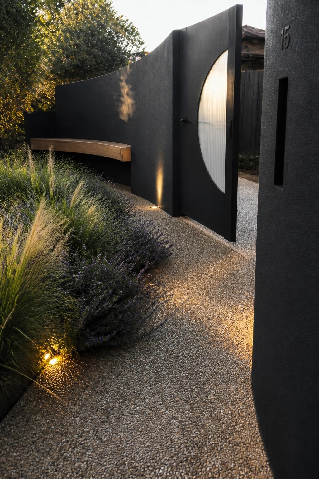 A black modern gate with a large circular ribbed panel stands open beside a curved black wall, gravel path, and garden plants with ground lighting.