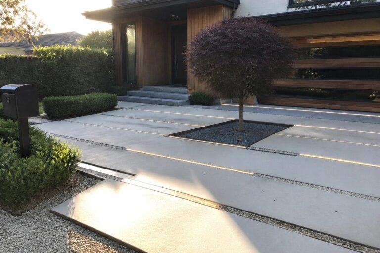 Modern concrete driveway pavers separated by gravel strips with embedded linear lights leading to a wood-clad house entrance and garage.