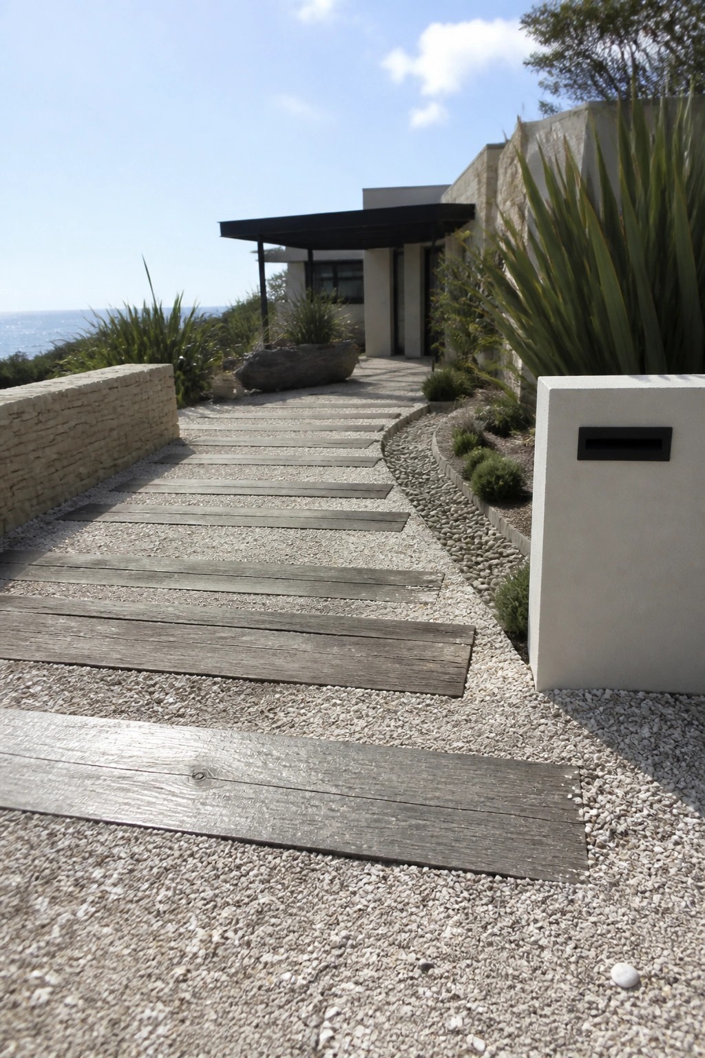 A gravel path with embedded wooden planks leads toward a modern house with stone walls and coastal plants.