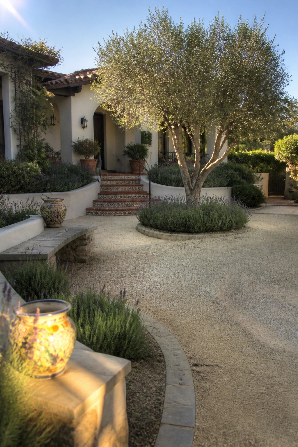 A gravel driveway curves past a large tree and landscaped beds toward the entrance of a stucco house with tiled steps.