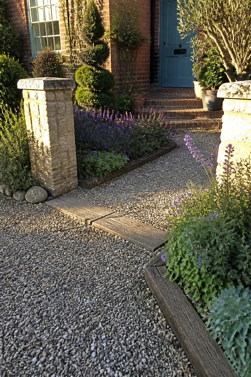 A gravel driveway bordered by wooden planks runs past stone pillars and purple flowering plants toward a brick house with a blue door.