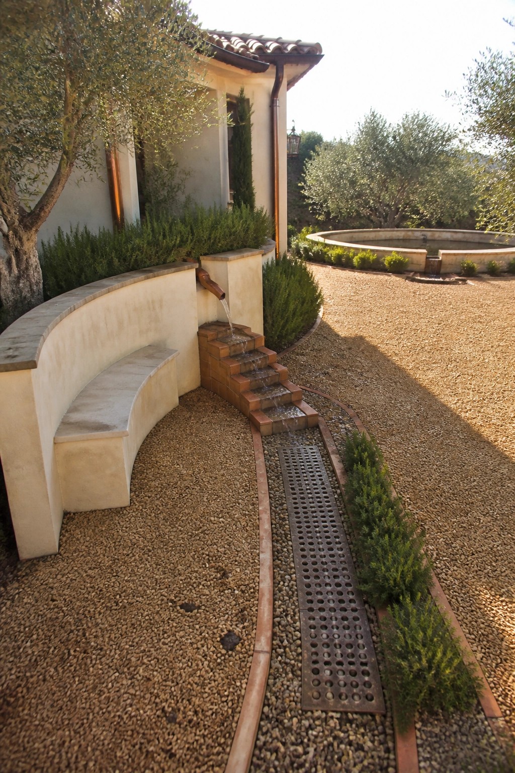 A gravel driveway features a long metal channel drain beside terraced brick steps with water flowing from a spout, next to a curved stucco wall and olive trees.