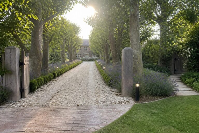 A long gravel driveway lined with tall trees leads toward a stone house, with low black path lights, lavender borders, wooden gates, and a brick section in the foreground.