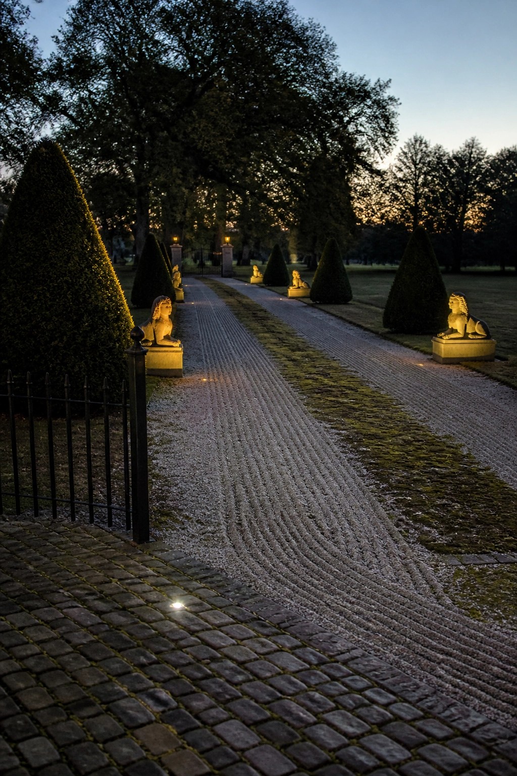 A long gravel driveway lined with sphinx statues on pedestals and conical hedges, viewed from a cobblestone edge at dusk.