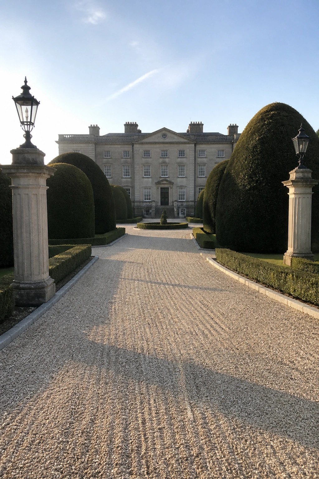 A long gravel driveway leads straight to a large stone mansion, bordered by tall clipped topiary bushes and stone pillars with lanterns.