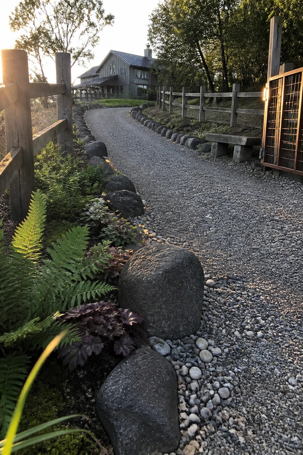 A gravel driveway curves past large dark rocks used as edging, with ferns and plants on one side and a wooden fence on the other.