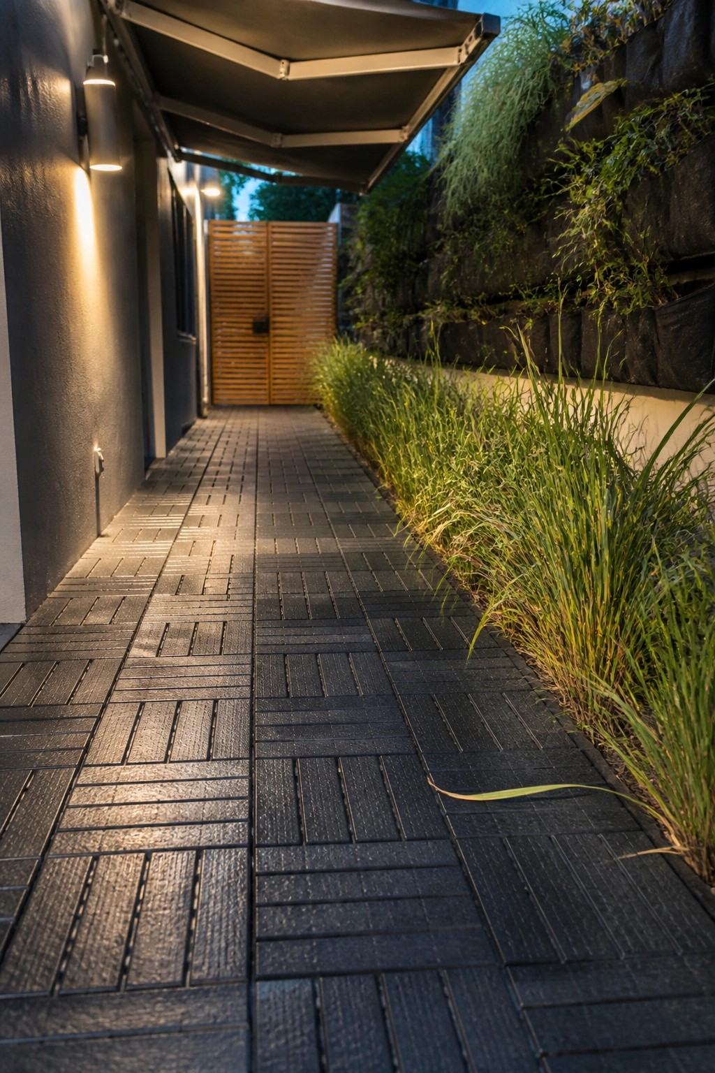 A narrow outdoor walkway covered in dark interlocking tiles, with tall ornamental grass along one edge and a stone retaining wall with greenery on the other.