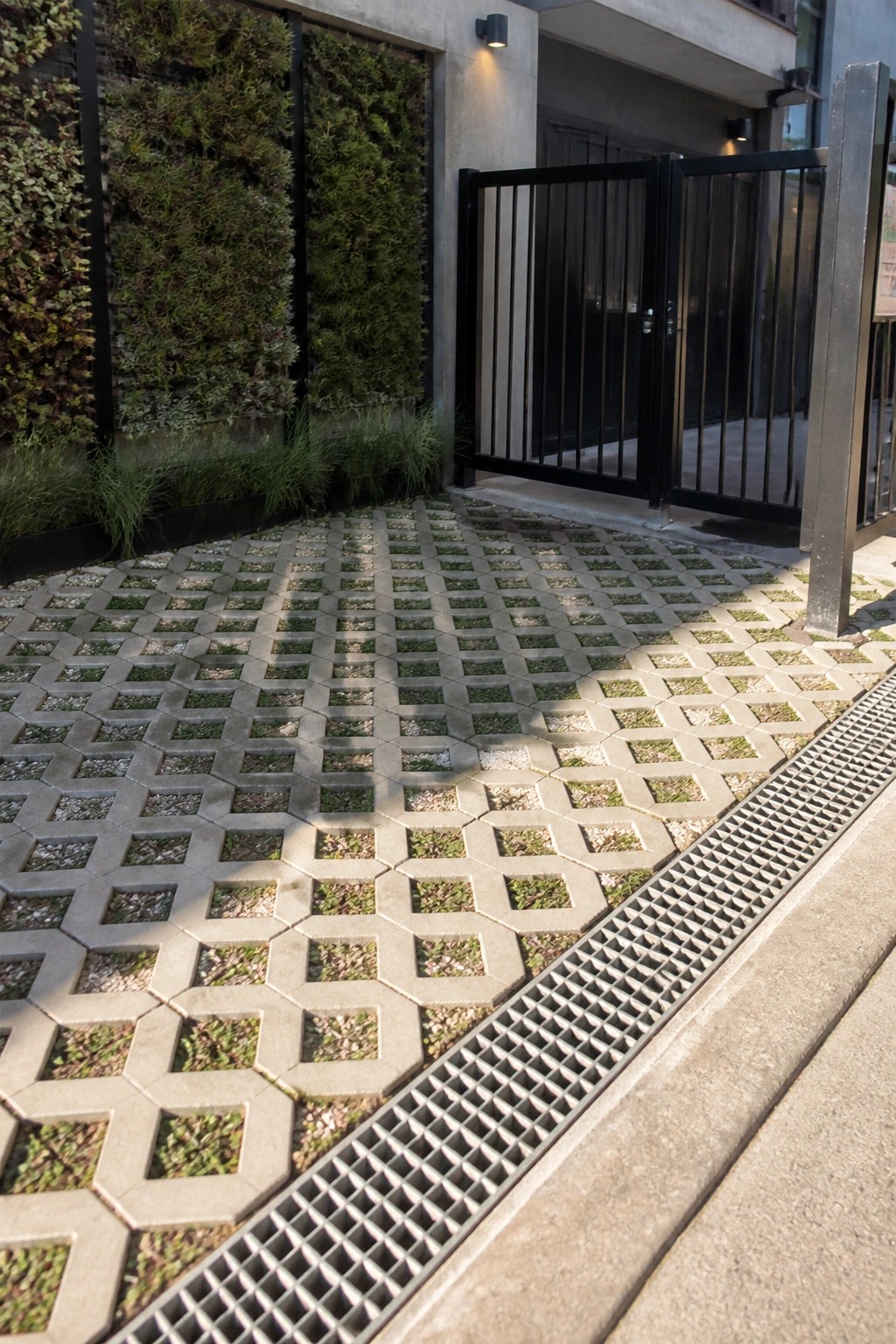 Hexagonal grass pavers with metal drain grate beside open black gate and green wall