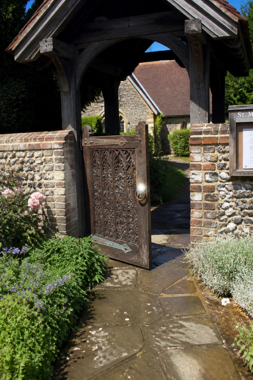Ornate carved wooden gate ajar in stone wall with flowers and church beyond