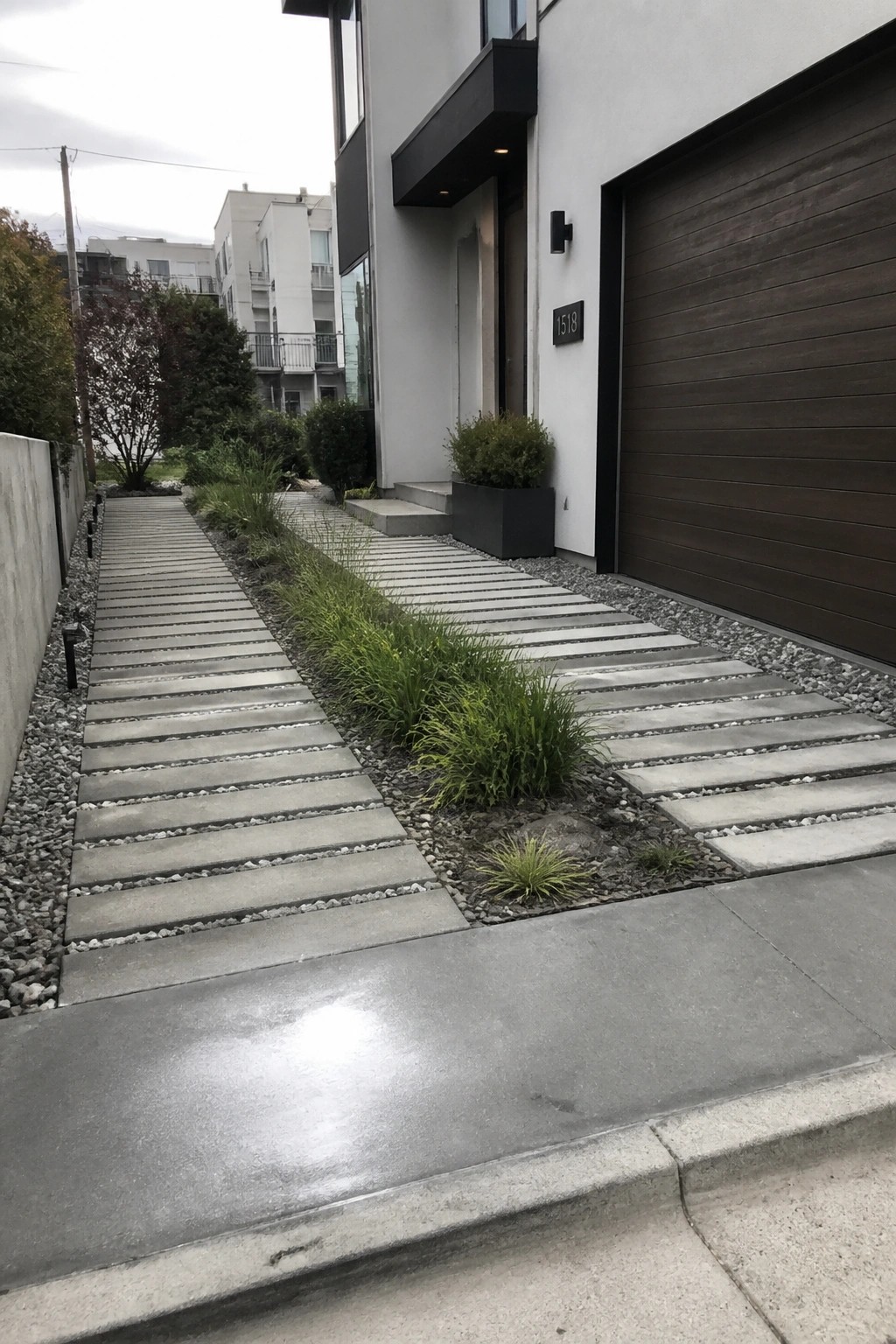 A modern house with a walkway of rectangular concrete pavers bordered by gravel and low green plants, next to a dark wood garage door.