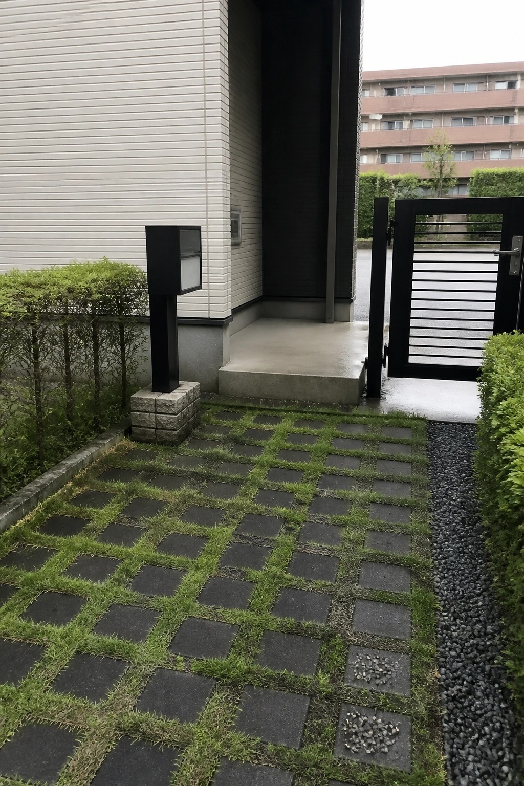 A grid of dark square pavers with grass growing between them forms a path leading to a house entrance beside a black metal gate.