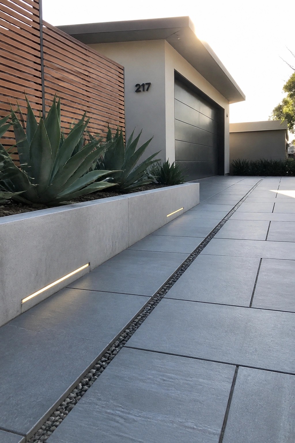 Modern house with a gray paver driveway that has a central strip of small gravel and a concrete planter with agave plants and linear lights.
