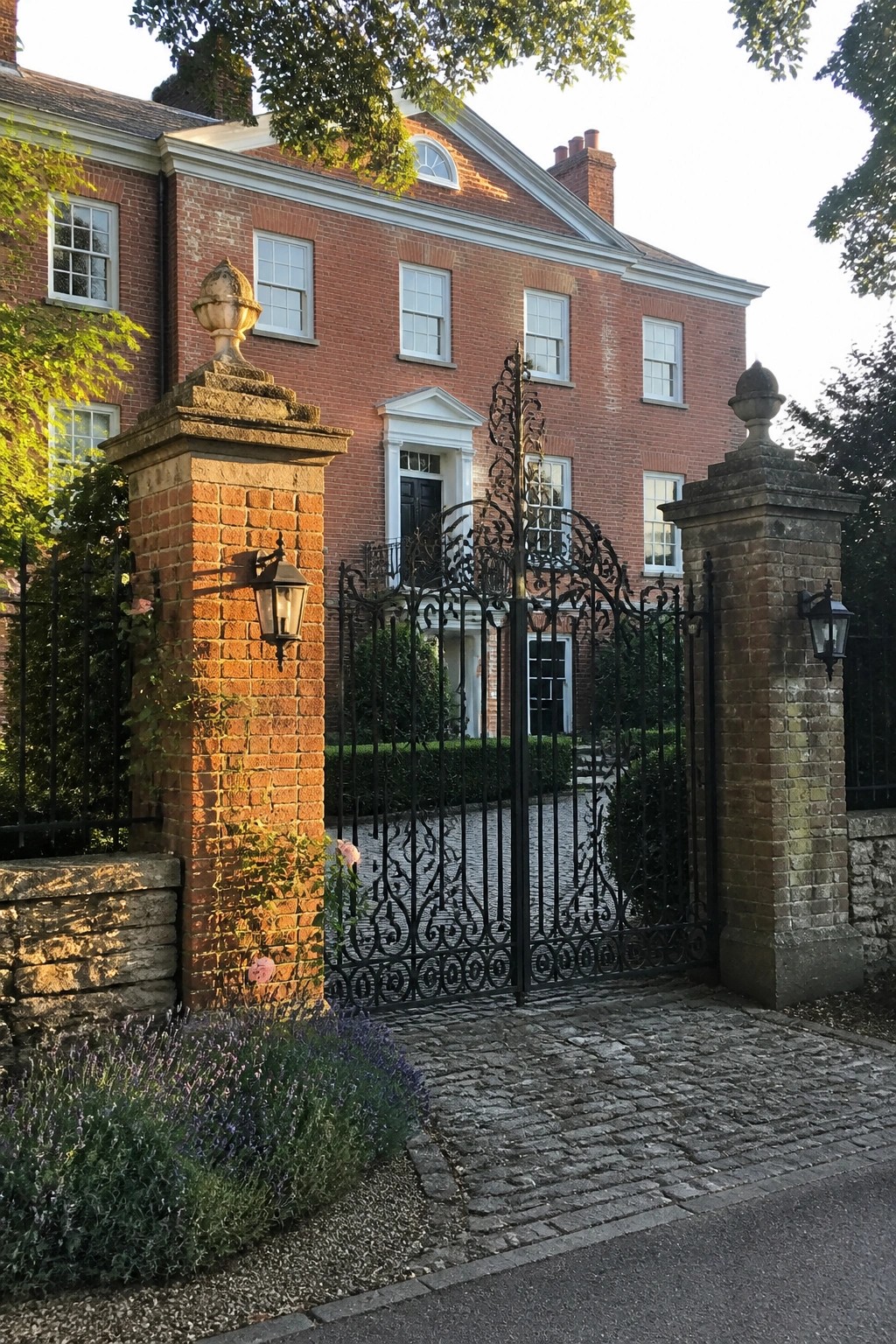 An ornate black wrought iron gate stands open between two brick pillars in front of a large red brick house with white trim.