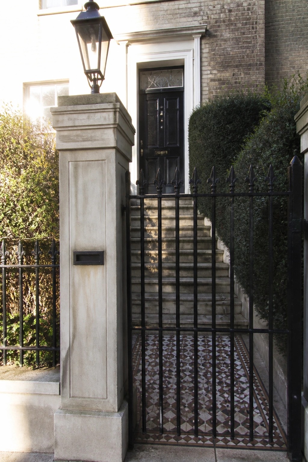 A black iron gate with pointed finials stands open beside a stone pillar in front of steps leading to a black door on a brick house.