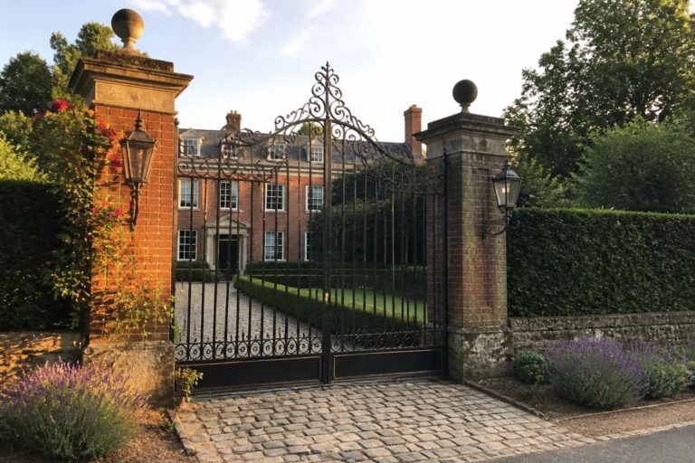 22 Classic Iron Main Gate Designs That Never Go Out of Style An ornate black wrought iron gate stands open between two brick pillars in front of a large red brick house with white trim.