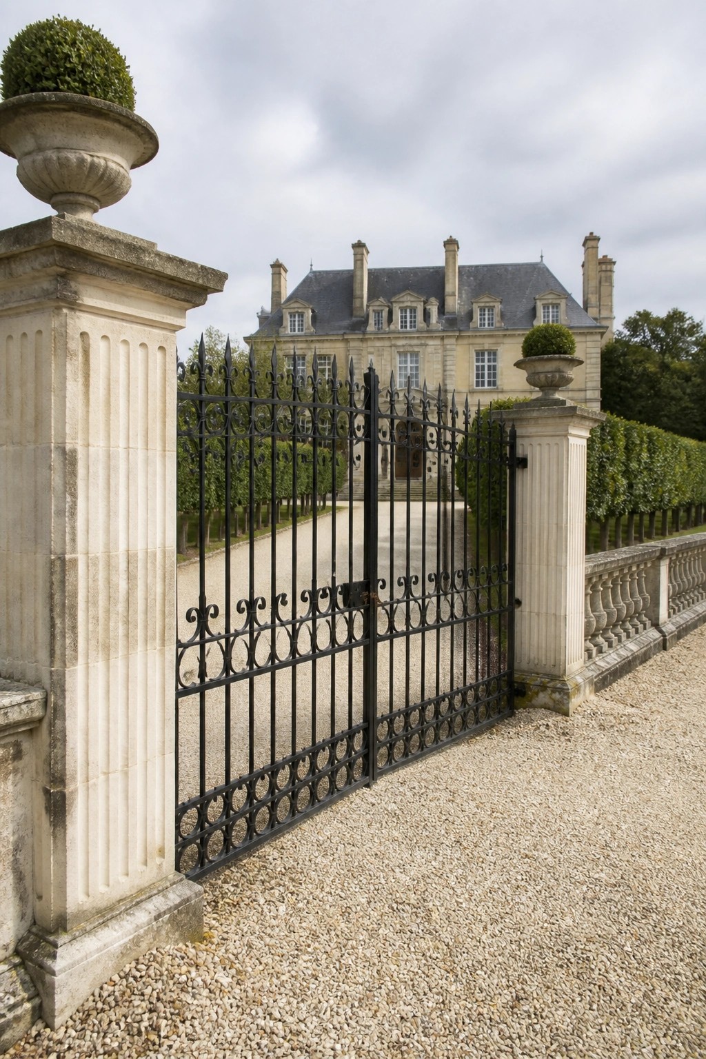 Ornate black wrought-iron gate between stone pillars with urns, chateau beyond gravel path.