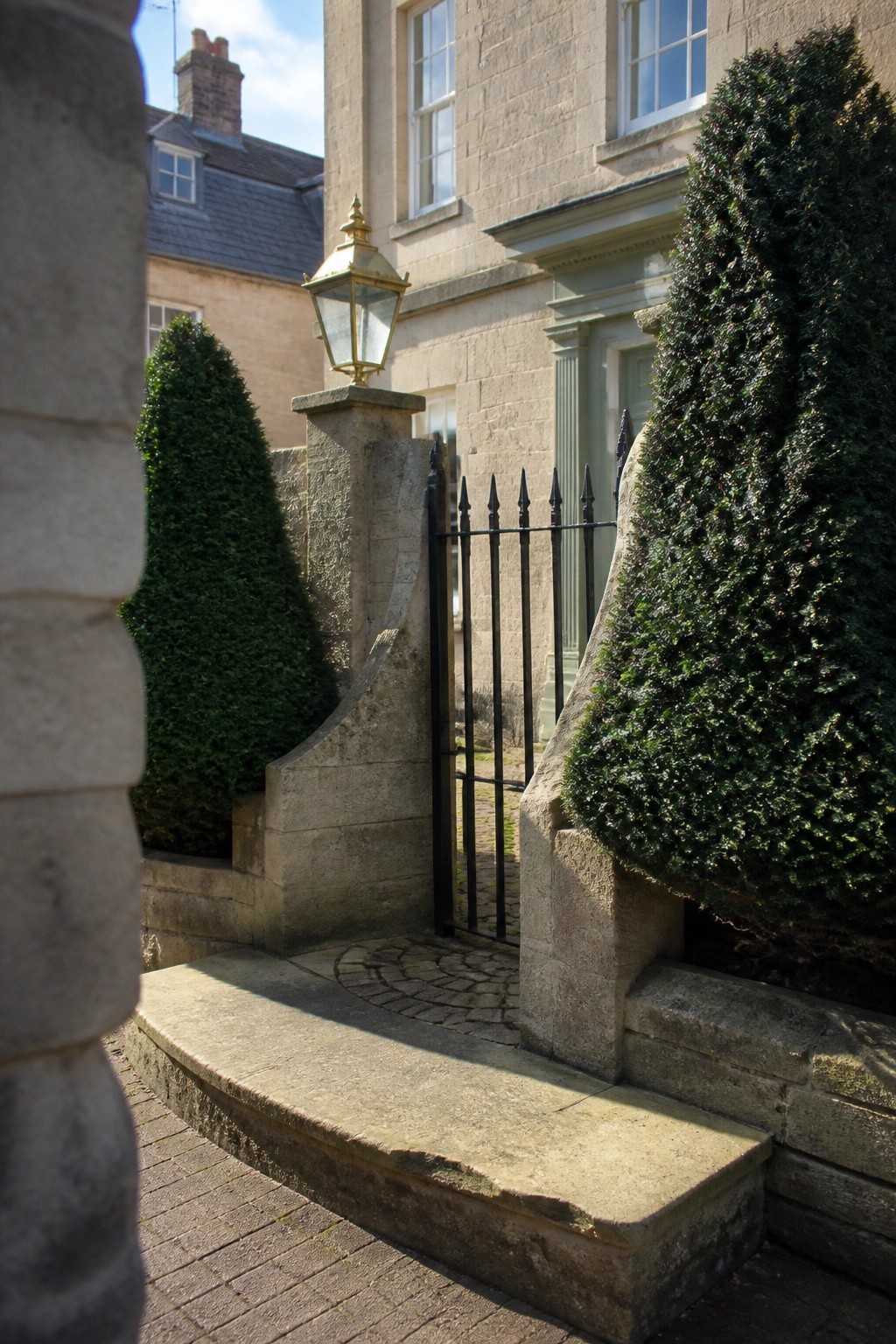 Stone entrance with black iron gate, golden lantern, and tall green shrubs