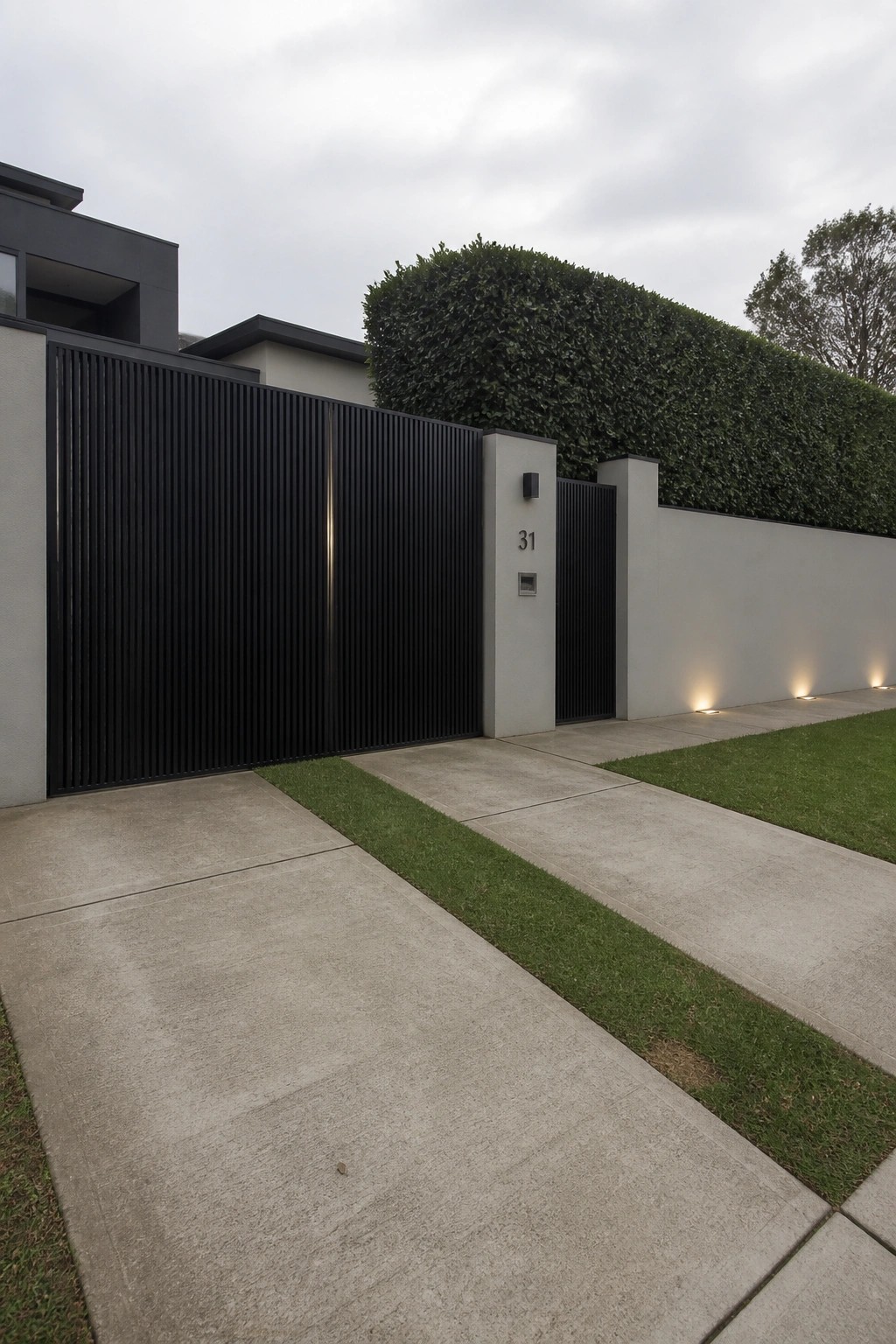Modern black slatted gate at house entrance with concrete driveway, white wall, and hedge.