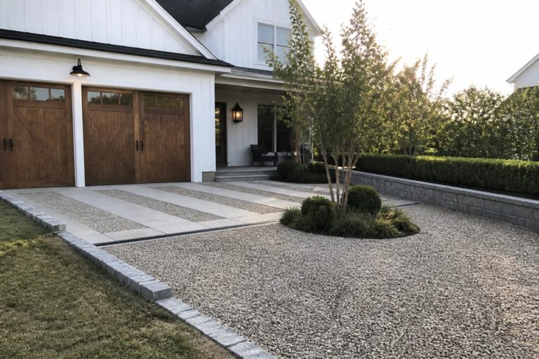 A gravel driveway with concrete paver strips, a long metal drainage grate, and stone borders leading to a white house with wooden garage doors.