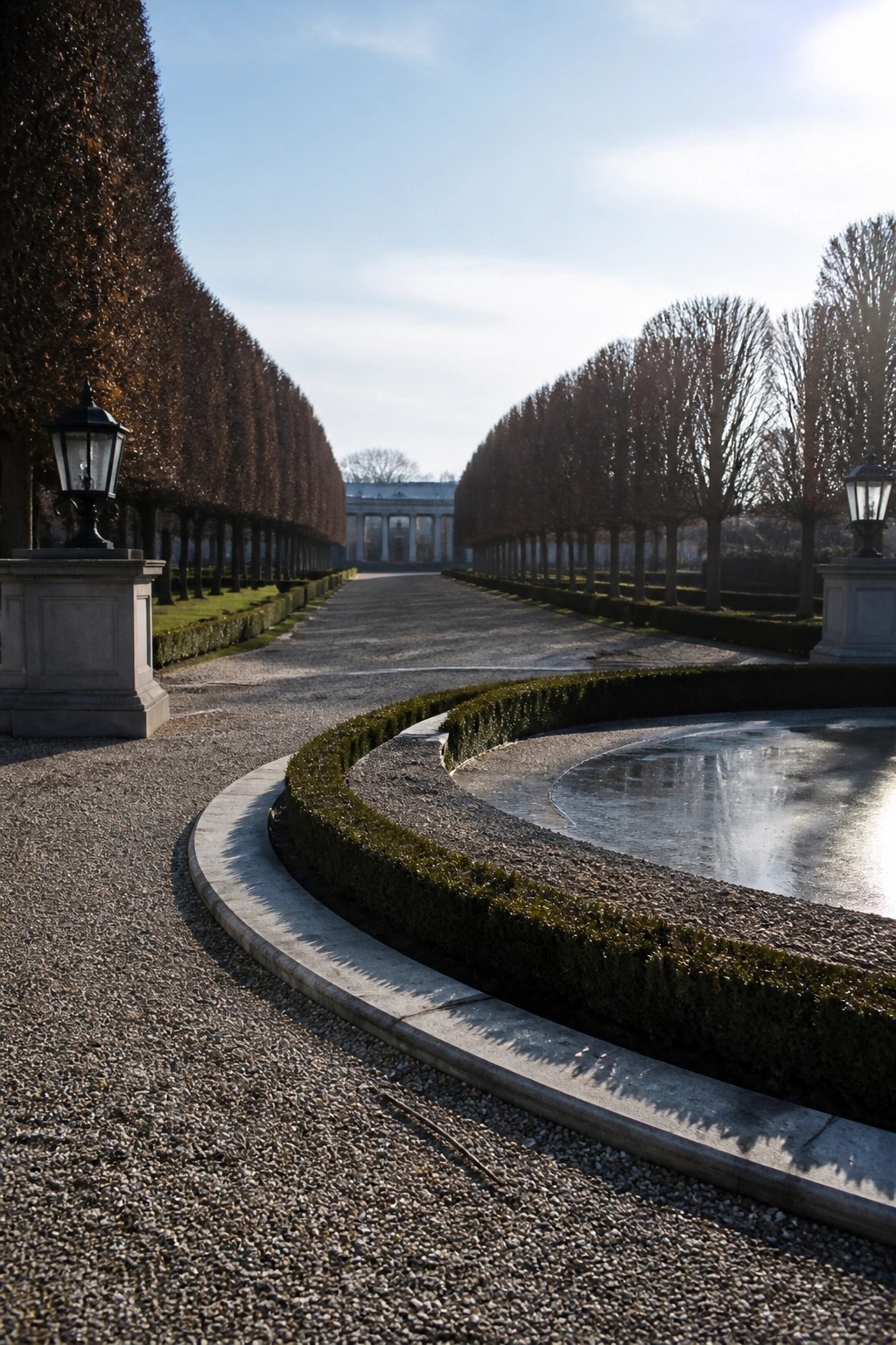 Curved gravel path beside frozen pond, flanked by hedges, lanterns, and bare trees.