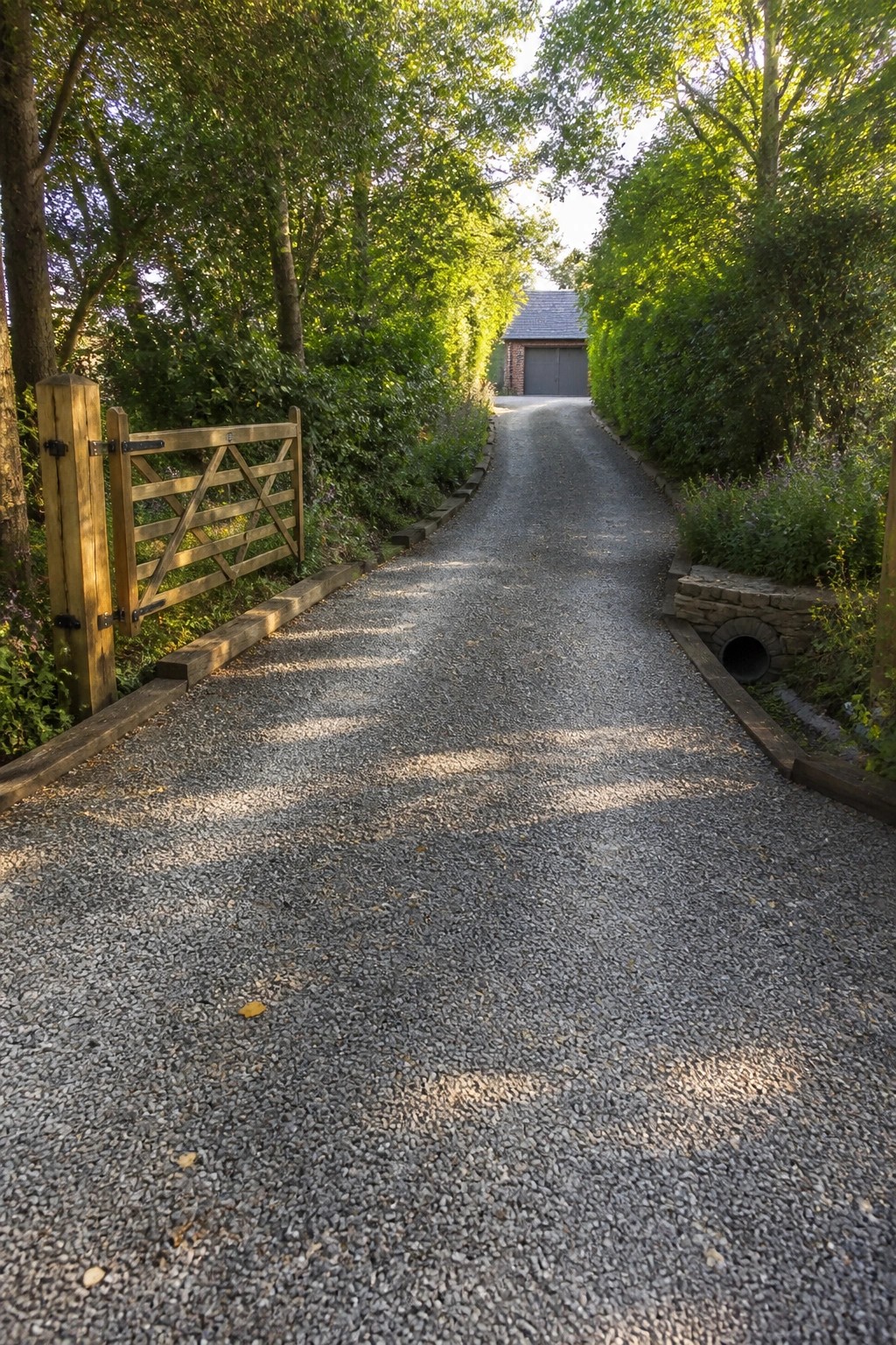 A gravel driveway bordered by wooden edging runs past a gate toward a distant building with trees on both sides.