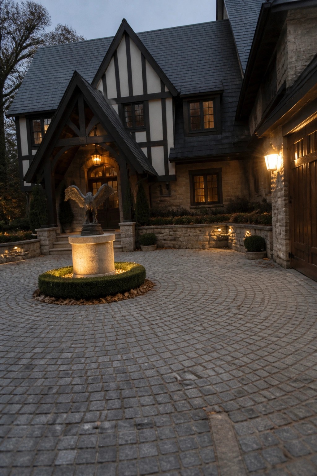 Tudor mansion with eagle fountain on lit cobblestone driveway at dusk