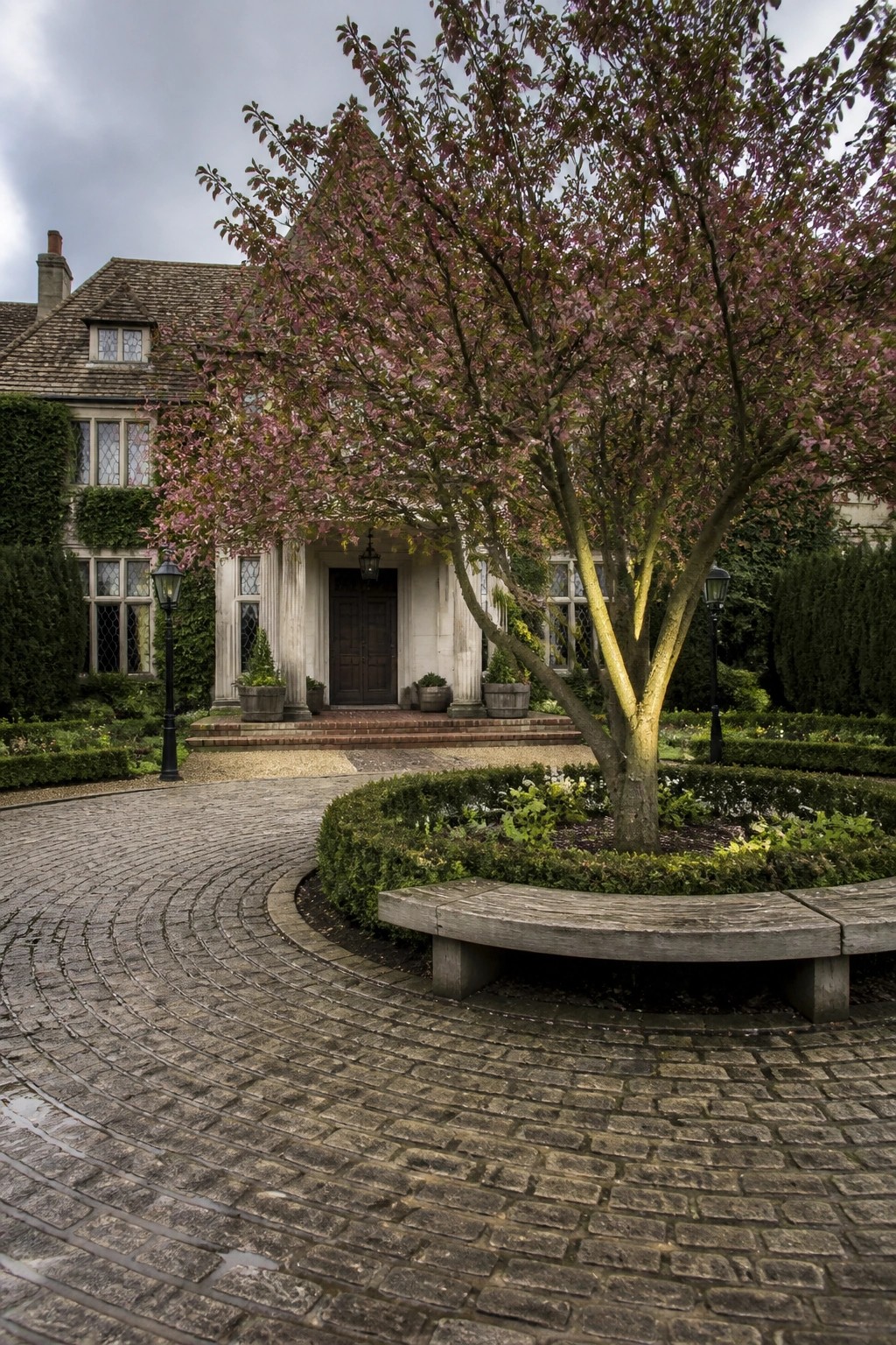 A large house with a cobblestone circular driveway surrounding a central tree and curved wooden bench, with potted plants near the entrance.