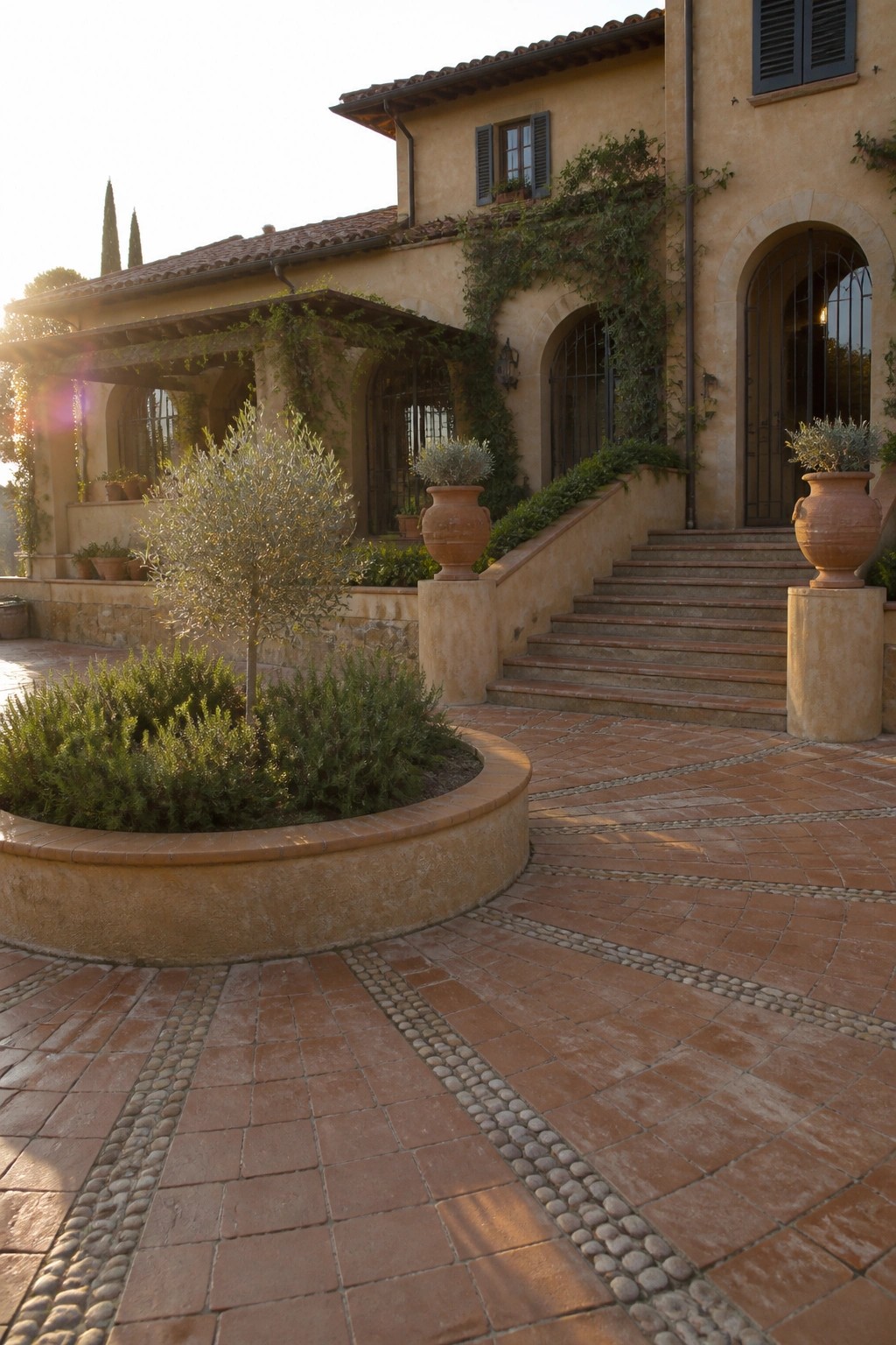 A terracotta tile circular driveway with pebble borders leads to a stucco house, featuring a central round garden bed with shrubs and a small tree, plus stairs flanked by large clay pots.