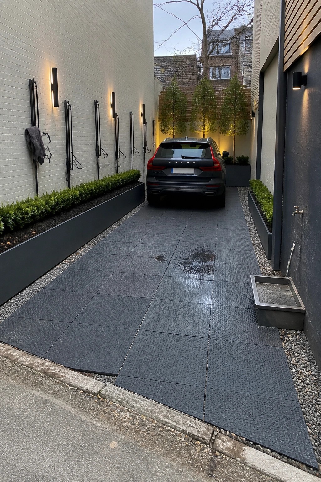 A narrow driveway lined with dark square tiles leads to a parked dark SUV, with white brick walls on the left, dark planters along both sides, and gravel borders.