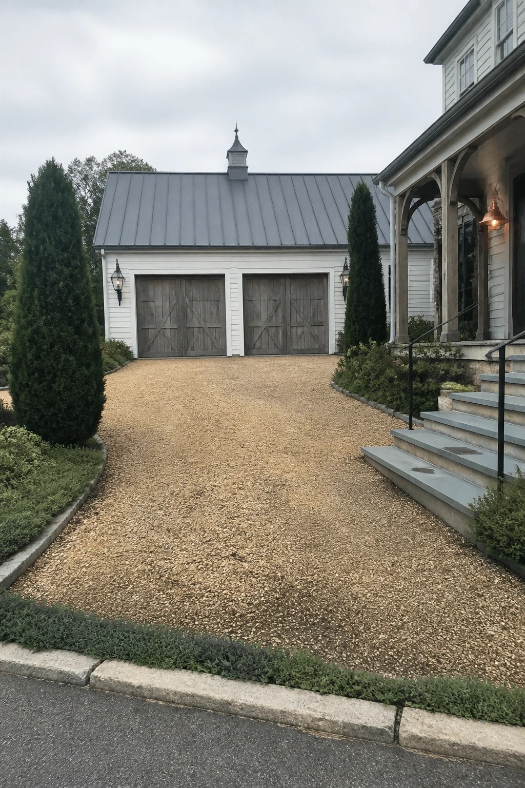A gravel driveway leads to a white garage with two sets of wooden barn doors, flanked by tall evergreen trees and a house porch with steps on the right.