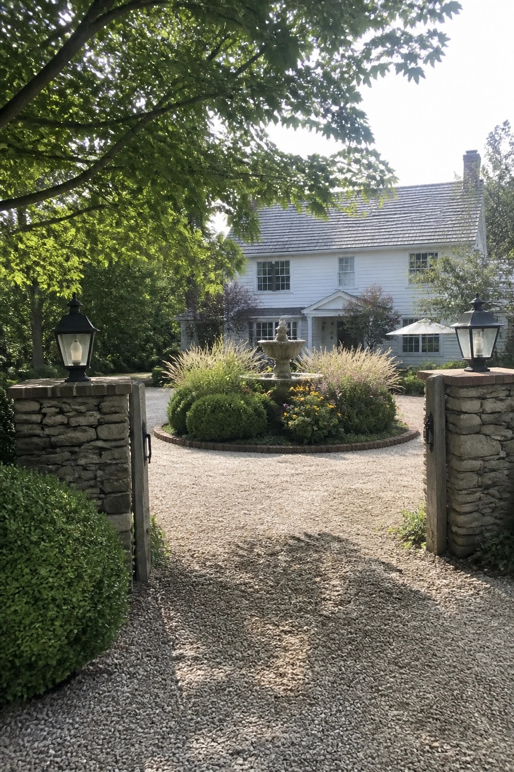 A gravel driveway passes through open wooden gates on stone pillars with black lanterns toward a white farmhouse, with a central circular garden bed and fountain in view.