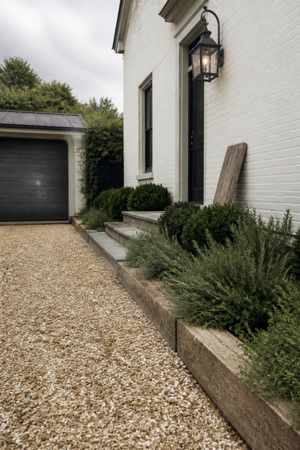A gravel driveway with wooden timber edging runs beside a white brick house, leading to concrete steps with green shrubs planted along the border.