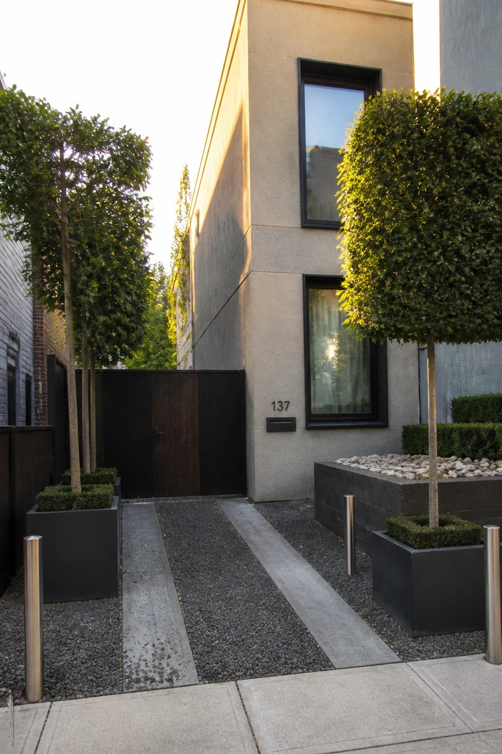 Modern concrete building with gravel path, potted trees, wooden gate, and number 137.