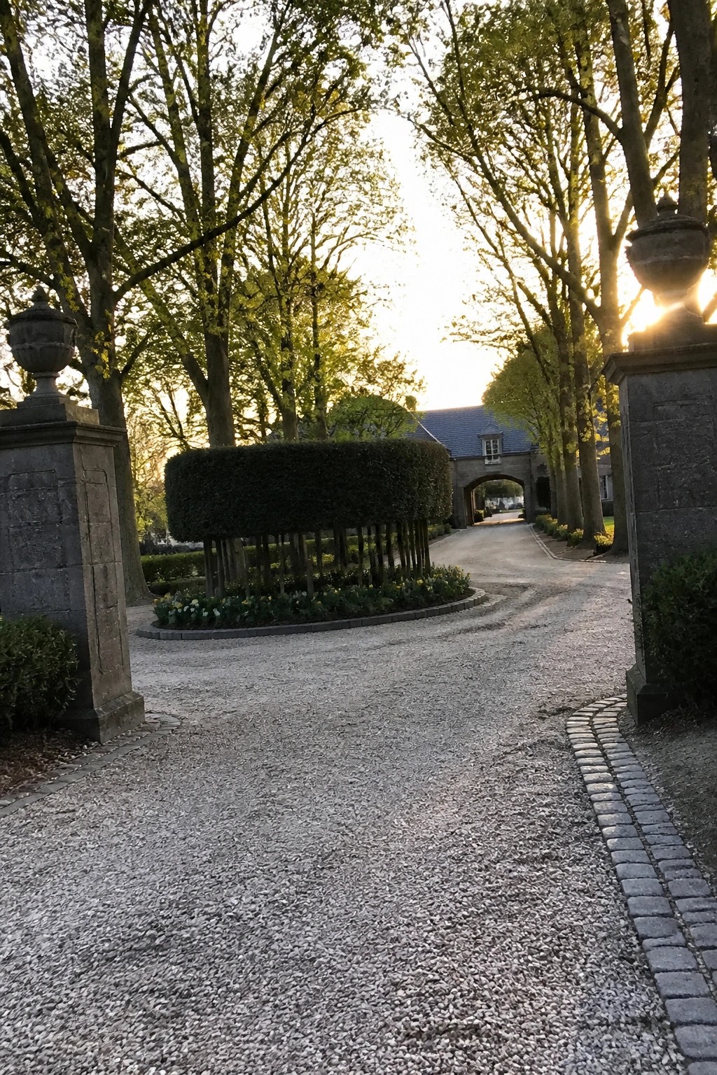 A gravel driveway curves between stone pillars past a round hedge and flower bed toward a house with an arched gateway.