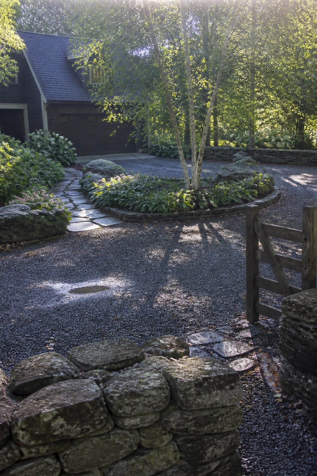 A gravel driveway curves in a circle around a central planting bed with trees and shrubs, bordered by low stone walls, in front of a dark house.