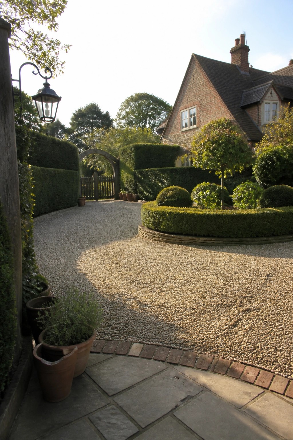 A curved gravel driveway bordered by low clipped hedges and brick edging leads toward a brick house with an arched gate.