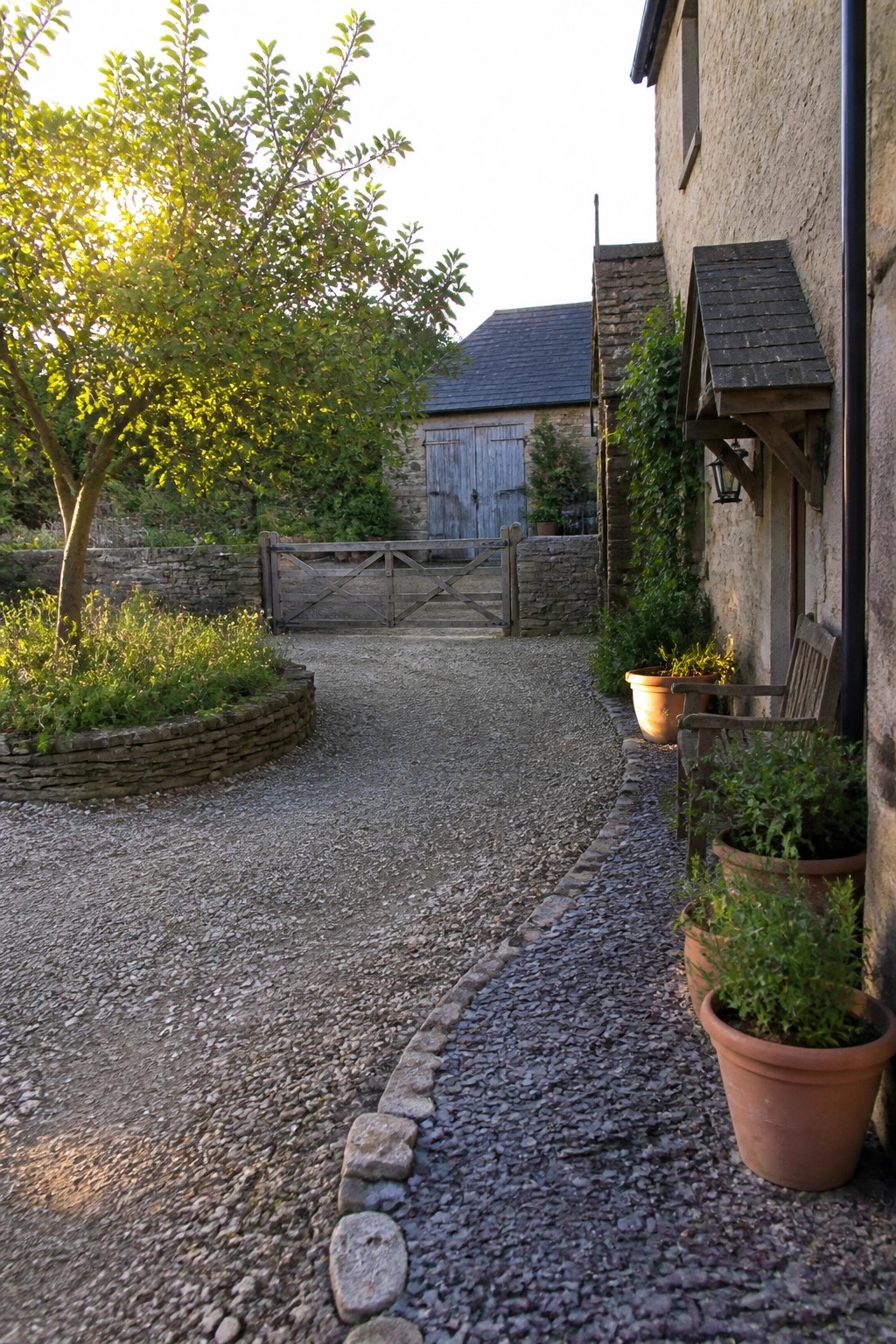 A gravel driveway curves past low stone edging and potted plants toward a wooden gate beside a stone building.