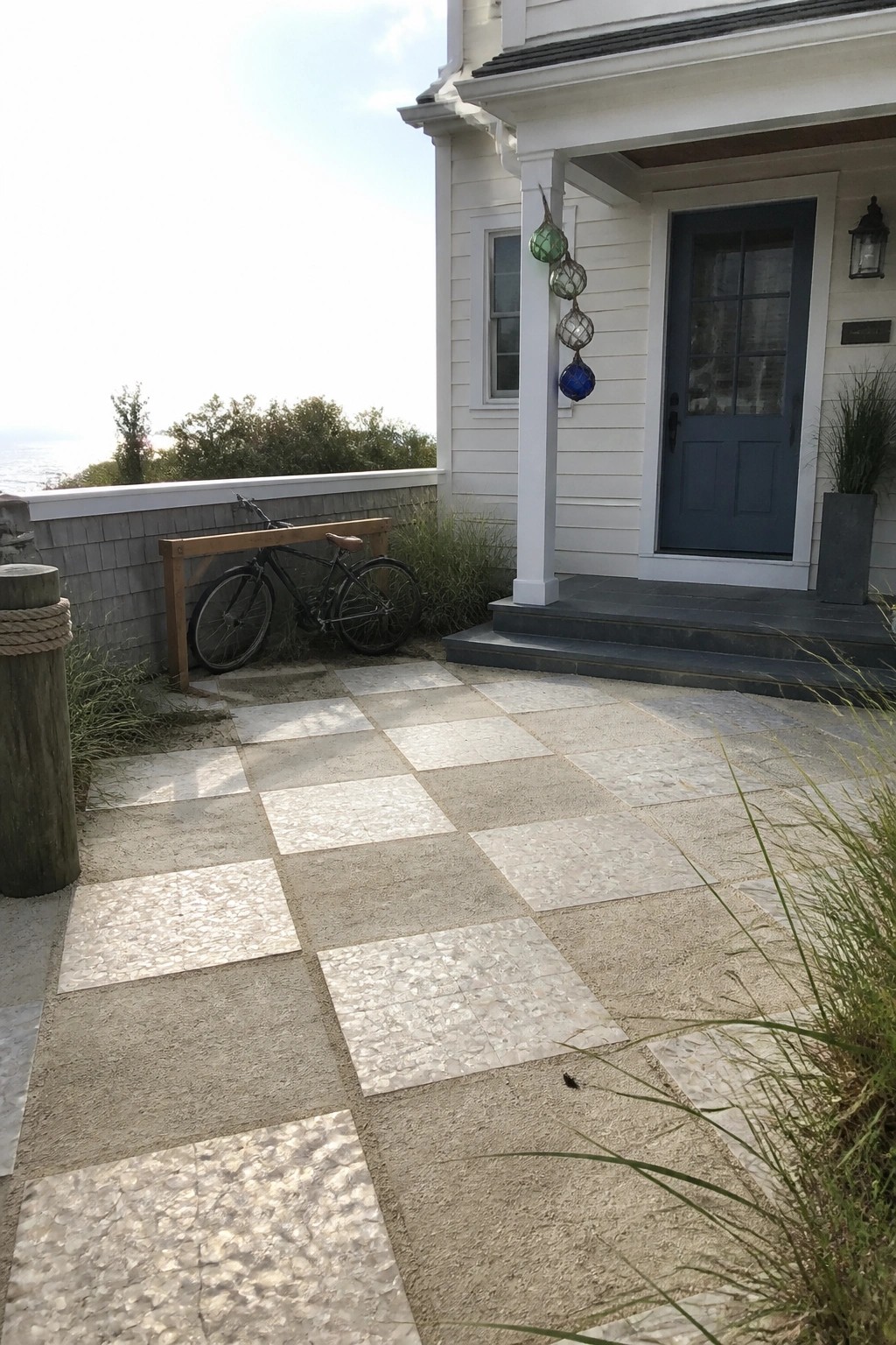 A checkered pattern of light and gray textured paving tiles leads up to the front steps of a white house with a blue door.