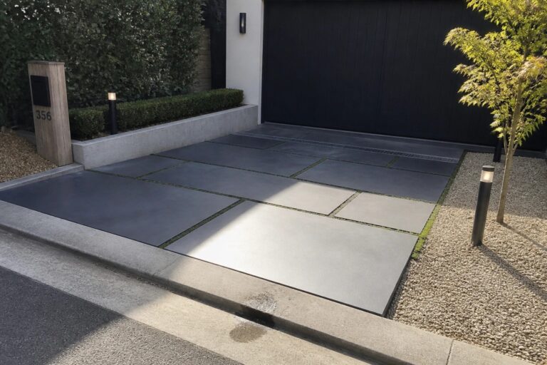 23 Creative Parking Tile Patterns That Transform Plain Spaces A modern driveway made of large gray rectangular pavers with narrow grass joints leading to a dark garage door, bordered by gravel beds and low shrubs.