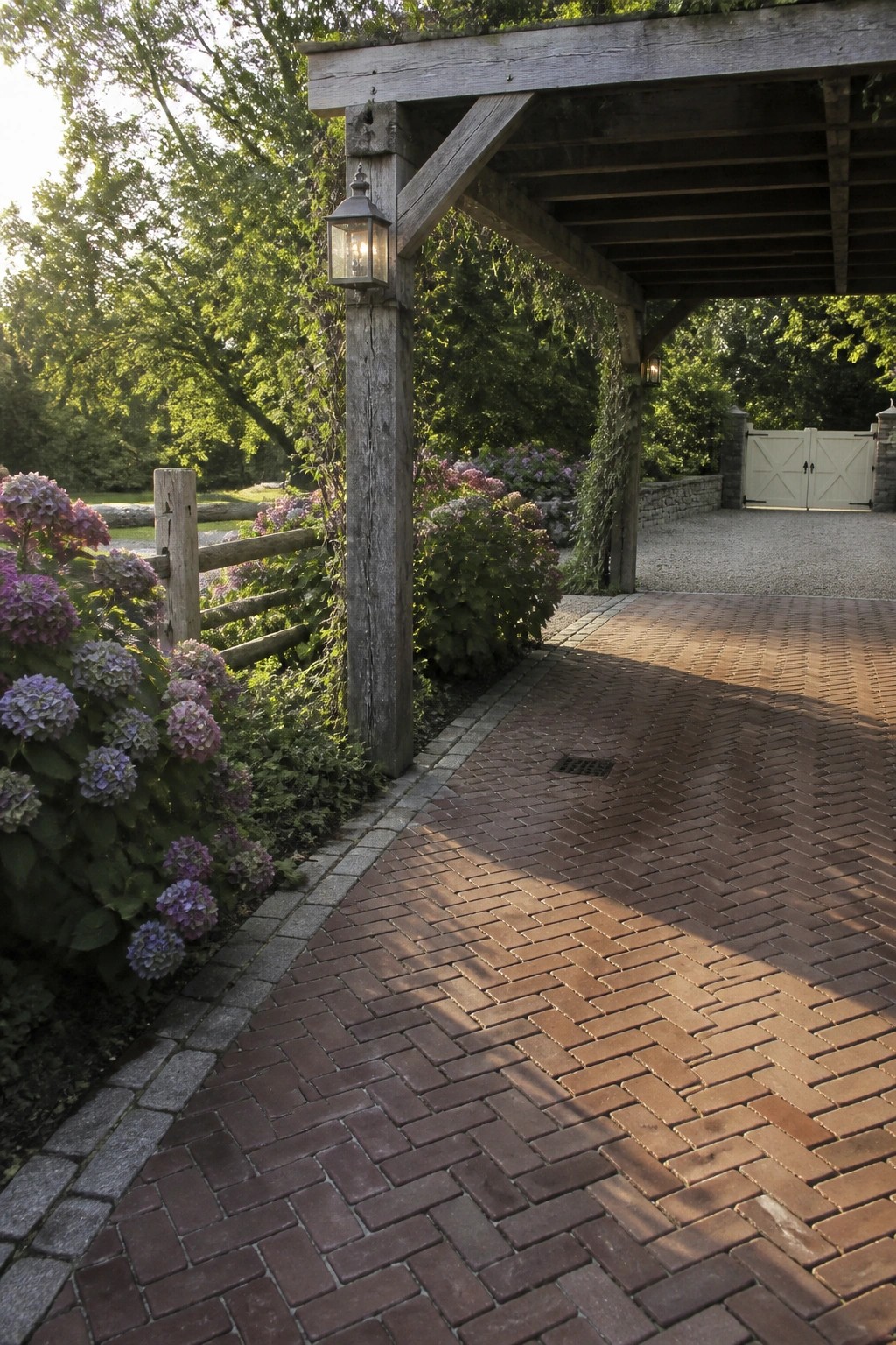 A herringbone brick driveway leads under a wooden carport, with hydrangeas and trees along the left side.