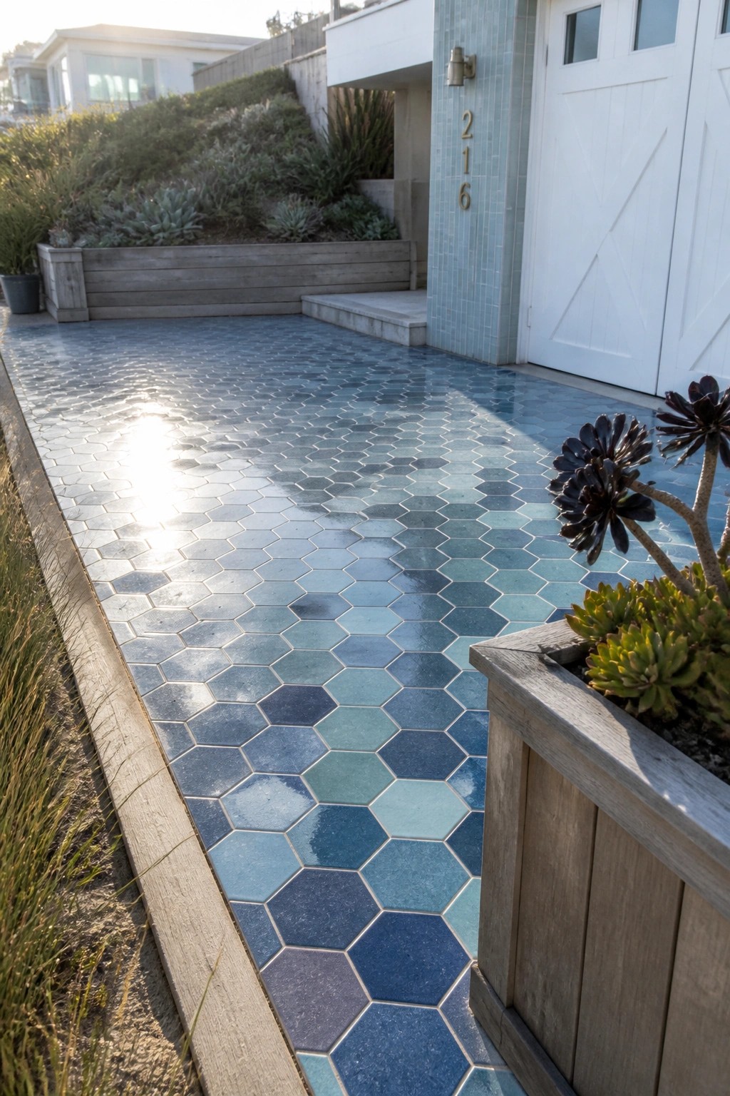 A driveway paved with hexagonal tiles in shades of blue and teal leads to a white garage door, with a wooden planter holding succulents visible on the right.