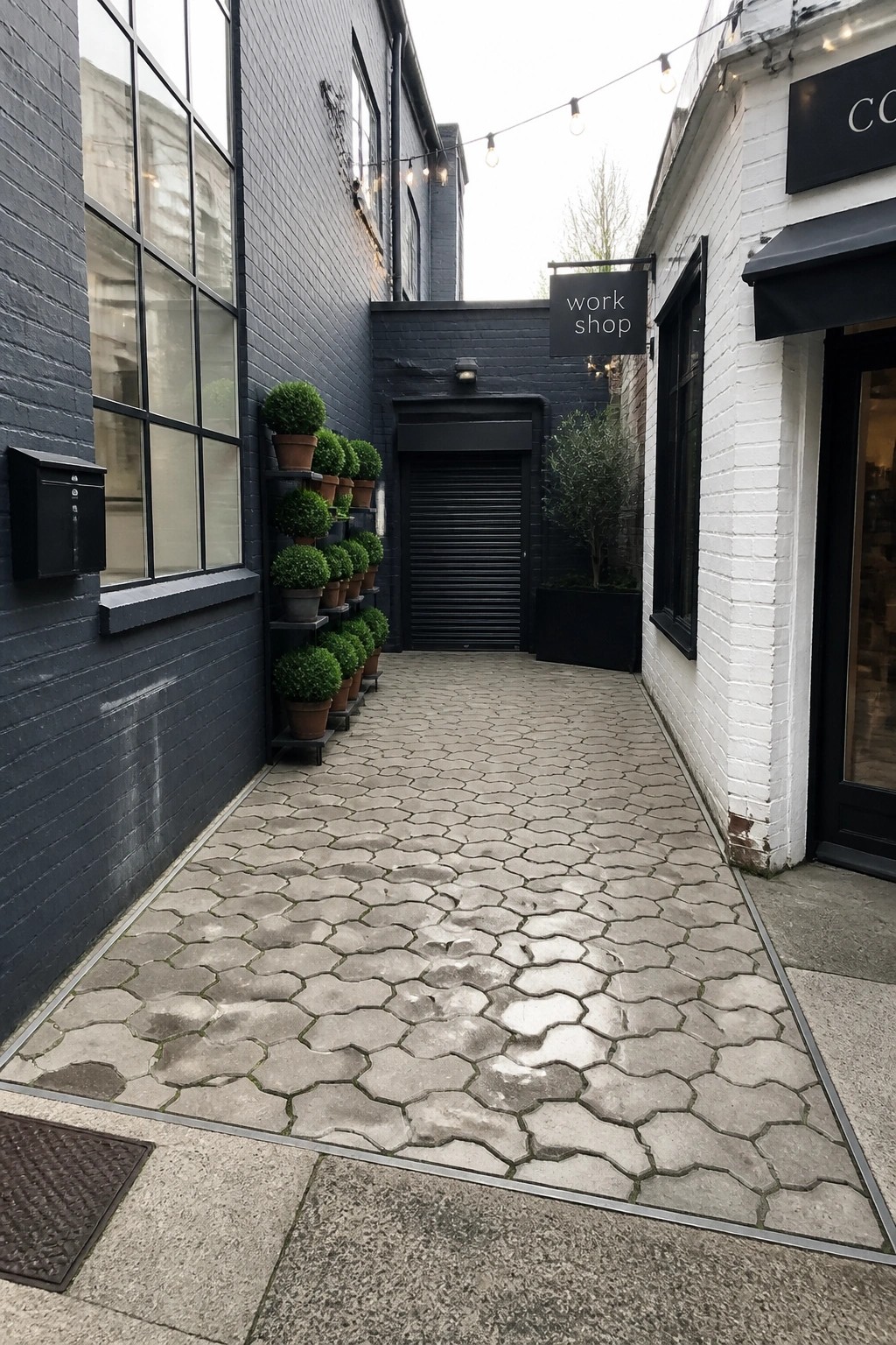 A narrow outdoor passageway paved with irregular light gray stone tiles between a dark brick building on the left and a white building on the right, with potted plants along one wall.
