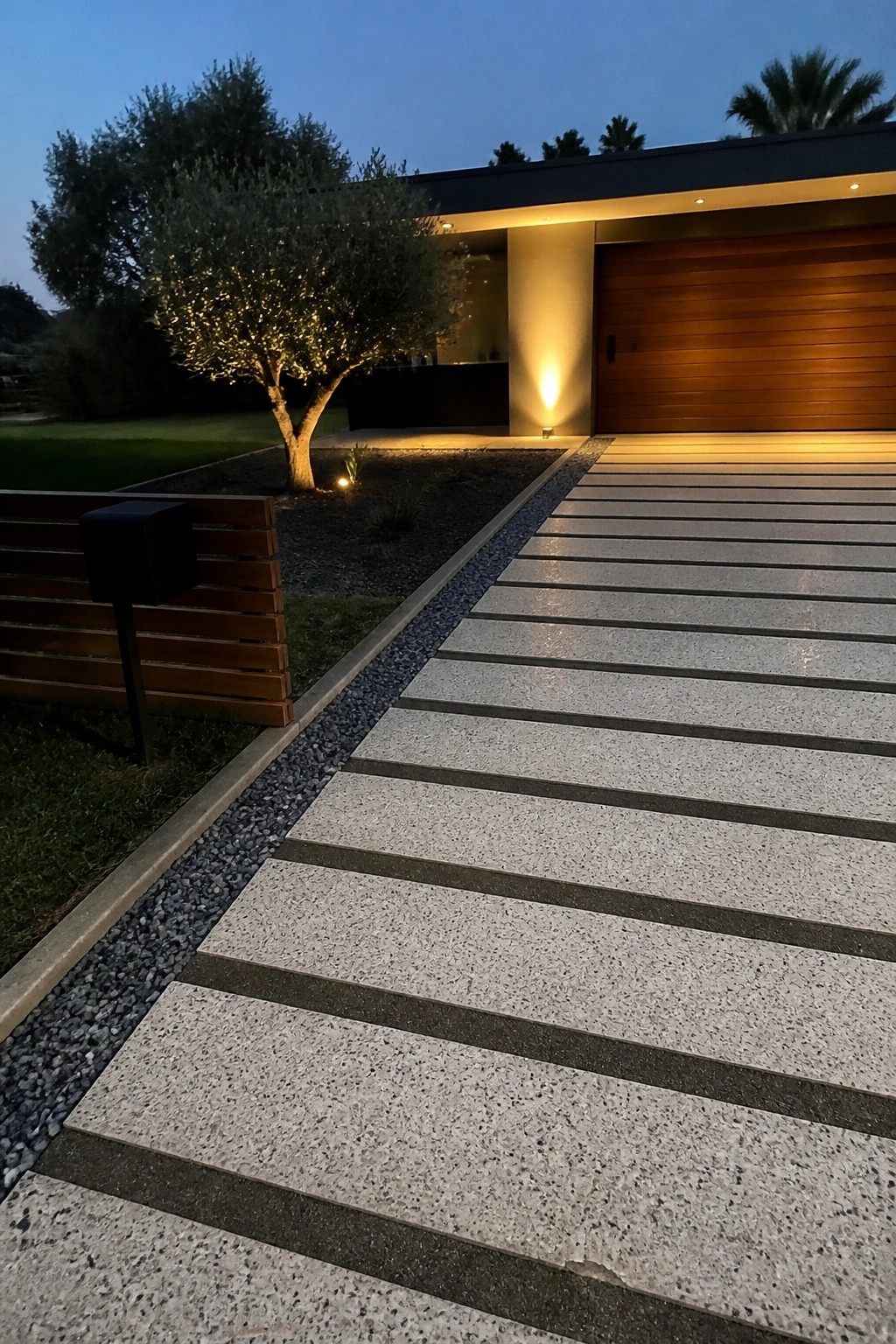 A modern driveway paved with light gray speckled concrete pavers separated by dark linear stripes, bordered by gravel and leading to a wooden garage door at dusk.