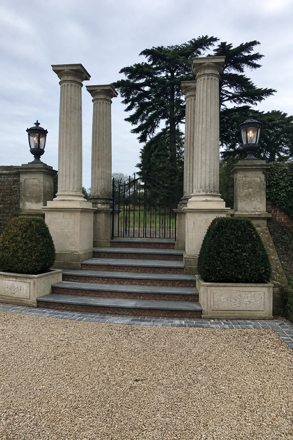 Tall fluted stone pillars stand on either side of a black metal gate, with lanterns on the outer pillars and steps leading through the entrance.