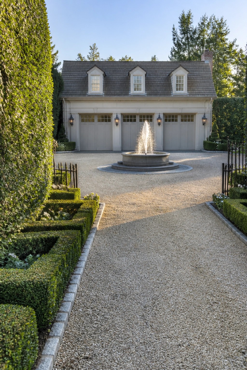 A gravel driveway with a round stone fountain in the center leads to a three-car garage with dormer windows and formal boxwood hedges on both sides.
